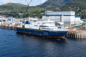 AN MV HUBBARD-LABELED SHIP IS DOCKED AT A PORT WITH SPRAWLING FOREST-FILLED MOUNTAINS IN THE BACKGROUND.