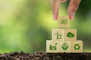 A HAND IS SETTING A BLOCK THAT READS "NET ZERO" ON TOP OF A SMALL PYRAMID OF OTHER BLOCKS THAT HAVE ESG-RELATED TERMS ON THEM. THE BLOCKS SIT ON A PATCH OF DIRT IN FRONT OF A GREEN BACKGROUND.