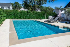 A PRISTINE RECTANGULAR SWIMMING POOL CAN BE SEEN IN A YARD FENCED IN BY TALL GREEN SHRUBS.