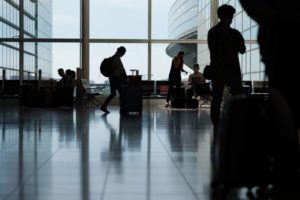 HUMAN FIGURES MOVE IN SILHOUETTE AGAINST THE GIANT GLASS WALLS OF AN AIRPORT TERMINAL