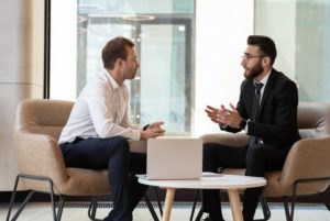 TWO MEN IN SUITS ENGAGE IN CONVERSATION ACROSS A COFFEE TABLE HOLDING AN OPEN LAPTOPIN AN OFFICE