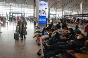  MASKED TRAVELERS PUSH SUITCASES THROUGH THE CONCOURSE OF AN AIRPORT, WITH SIGNAGE IN CHINESE