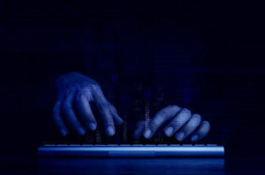 Digital binary code, data numbers and secure lock icons can be seen on a hacker's hands above a computer keyboard with a dark blue tone background. Photo: iStock.com/Techa Tungateja