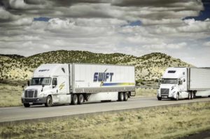 TWO TRUCKS BEARING THE KNIGHT-SWIFT LOGO DRIVE DOWN A HIGHWAY THROUGH DESERT