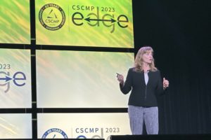 A WOMAN STANDS TALKING ON STAGE IN FRONT OF A CSCMP EDGE LOGO