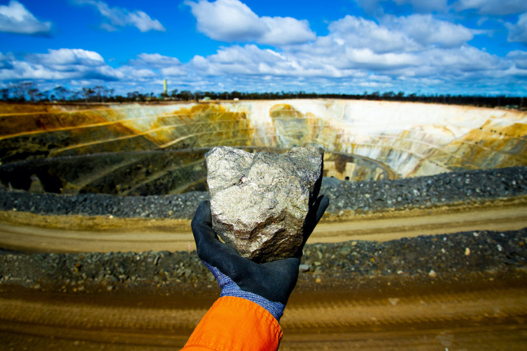 A PERSON WEARING A JUMP SUIT AND A GLOVE HOLDS A NICKEL ORE IN THEIR HAND IN FRONT OF A LARGE MINE.