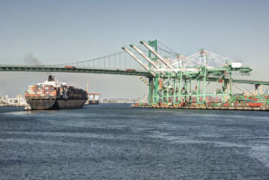 A LARGE CONTAINER SHIP SAILS UNDERNEATH A BRIDGE NEAR SOME CRANES AT A BUSY PORT.