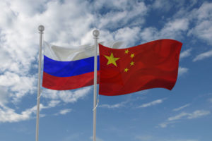 A RUSSIAN AND CHINESE FLAG WAVE NEXT TO EACH OTHER ON FLAG POLES IN FRONT OF A BLUE, CLOUDY SKY.