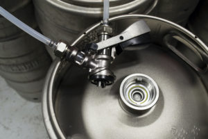 THE TOP VIEW OF A STEEL KEG OF BEER CAN BE SEEN WITH A COUPLER AND PIPES DETACHED FROM IT. OTHER KEGS CAN BE SEEN BEHIND IT.