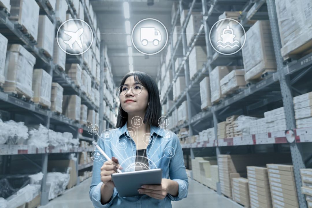 A WOMAN STANDS WITH A TABLET COMPUTER IN A WAREHOUSE, SURROUNDED BY SYMBOLS FOR SHIPPING METHODS