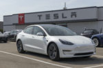 A WHITE TESLA VEHICLE SITS IN A PARKING LOT IN FRONT OF A BUILDING WITH THE TESLA LOGO ON IT.