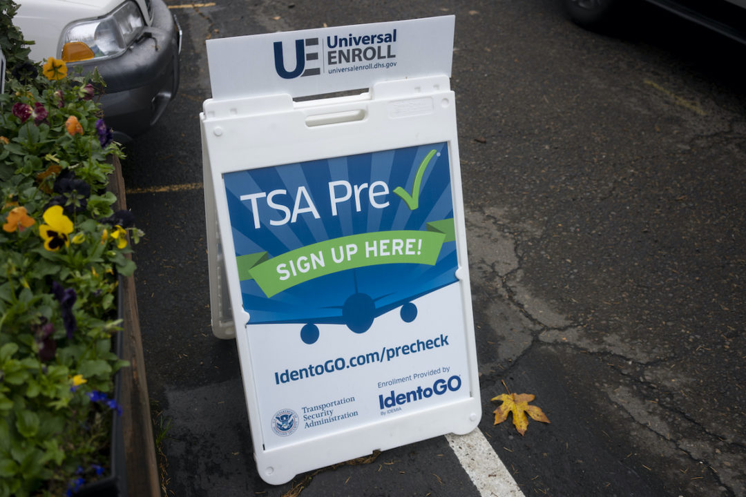 A SANDWICH BOARD SIGN ADVERTISING TSA PRE-CHECKS SITS ON A SIDEWALK NEXT TO FLOWERS.