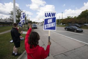 A LINE OF PEOPLE BY THE SIDE OF A ROAD HOLD UP SIGNS SAYING UAW ON STRIKE TO PASSING CARS
